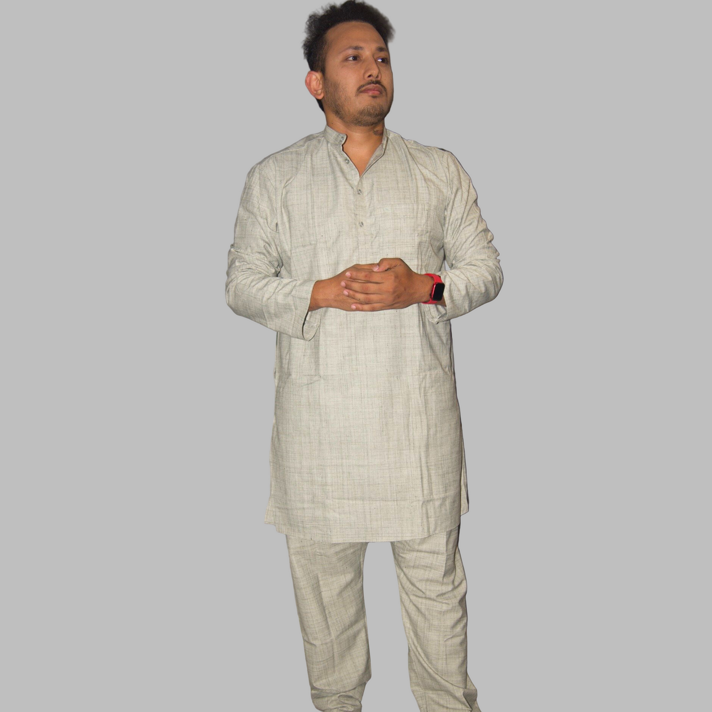 Man wearing a light gray traditional outfit standing against a plain wall.