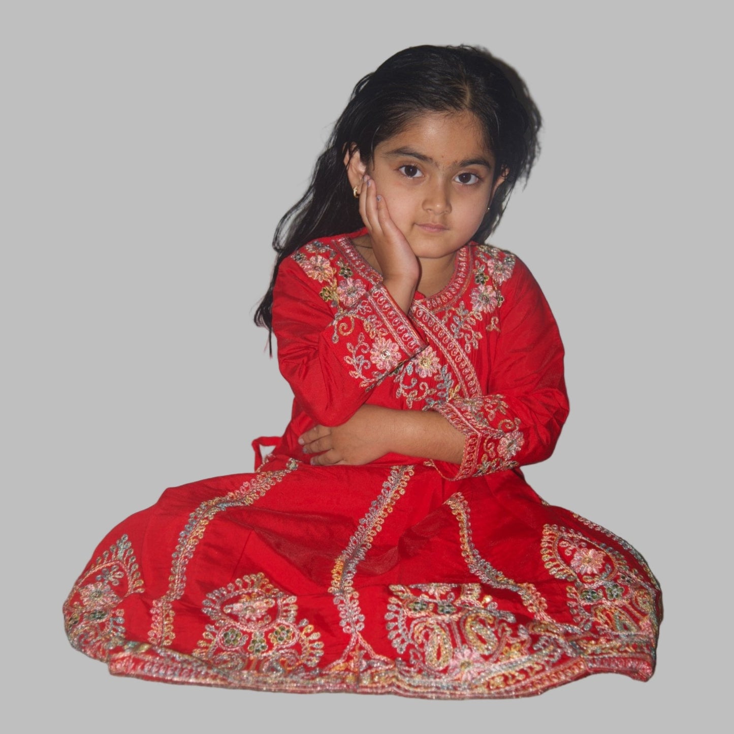 Young girl in a red traditional outfit sitting on the floor against a plain wall.