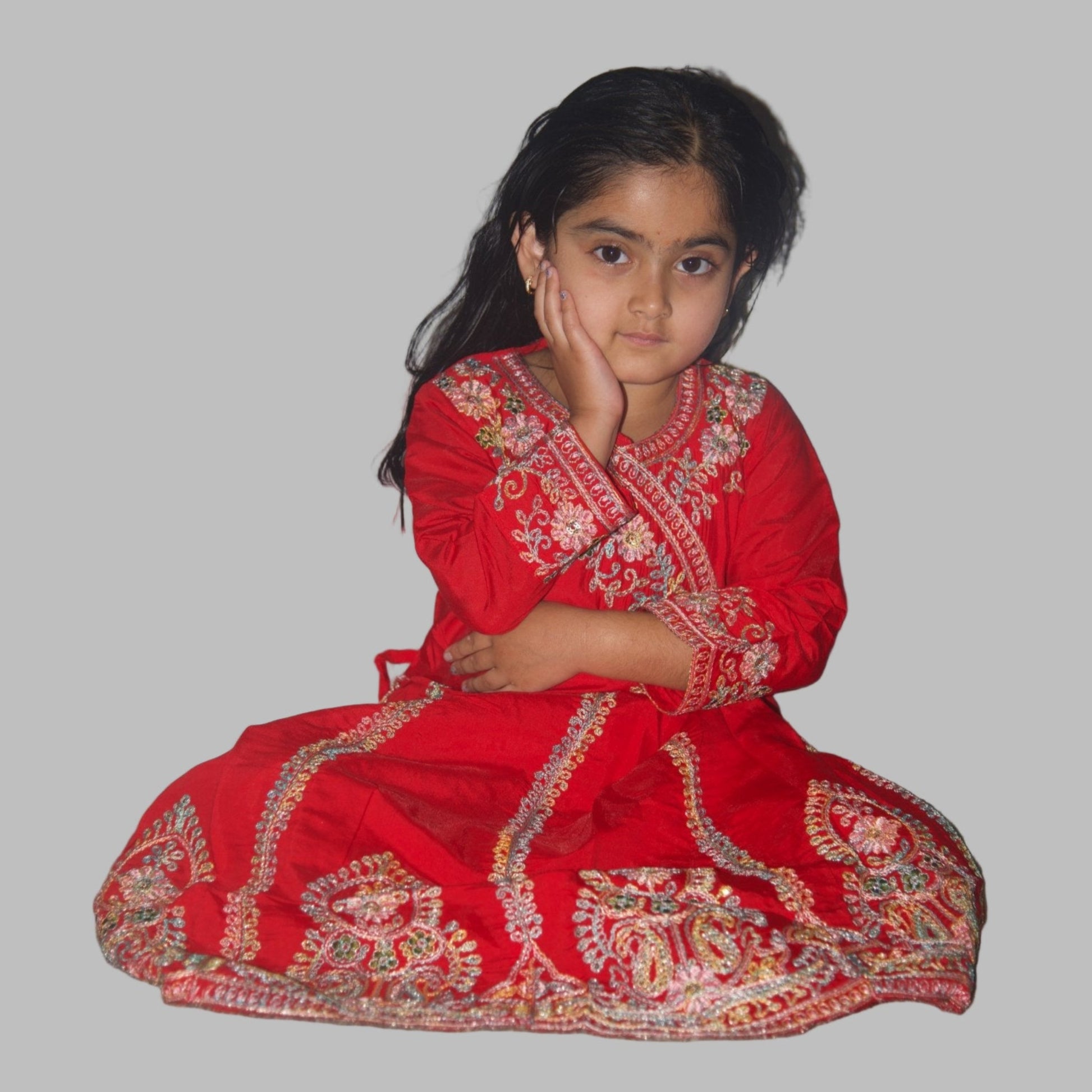 Young girl in a red traditional outfit sitting on the floor against a plain wall.