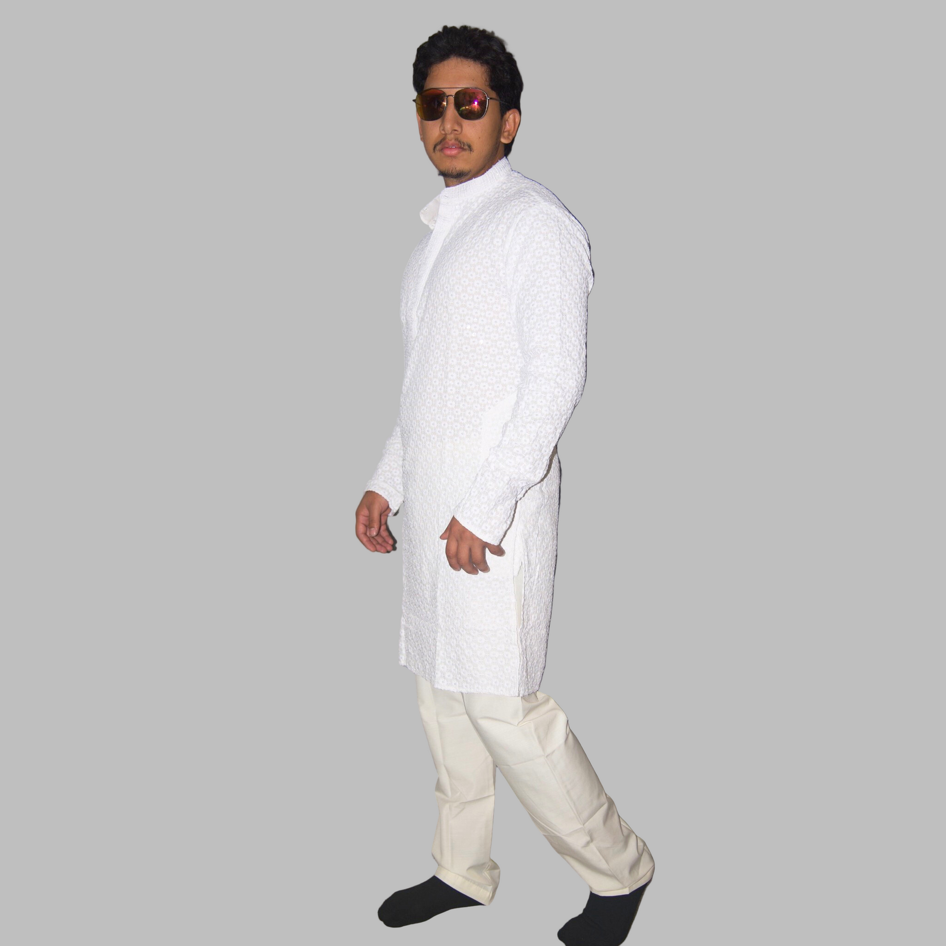 Man wearing a white kurta with black shoes against a plain wall.