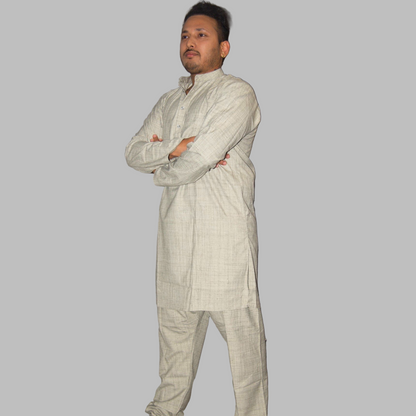 Man wearing a light-colored traditional outfit standing against a plain wall.